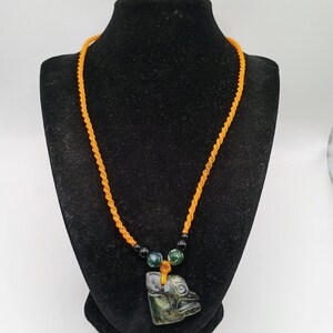 May include: An orange cord necklace with a carved green pendant in the shape of a bird's head. The necklace has green and black beads near the pendant. The necklace is displayed on a black mannequin.