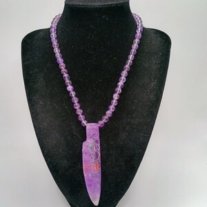 May include: A necklace featuring a strand of round, translucent purple amethyst beads and a large, carved amethyst pendant shaped like a knife. The pendant includes small, inlaid circular accents in green and orange on its surface.