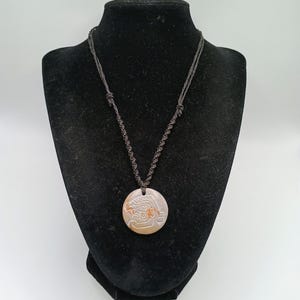 May include: A round pendant necklace with a carved design, displayed on a black mannequin. The pendant is tan and brown with a detailed relief carving. The necklace cord is dark brown, knotted, and hangs from the pendant.