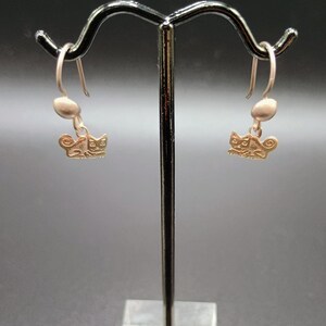 May include: A pair of gold-colored cat earrings. Each earring features a small, stylized cat charm hanging below a small, oval-shaped bead. The earrings are displayed on a silver-colored earring stand against a dark background.