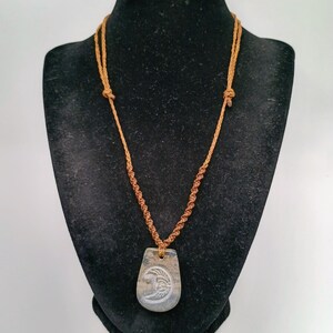 May include: A necklace featuring a brown cord with a twisted macrame design and a flat, teardrop-shaped pendant. The pendant is grey with a white crescent moon graphic etched into the center, suspended from the cord by a small loop at the top.