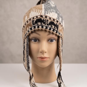 May include: A knitted earflap hat featuring a multi-coloured geometric and animal pattern in shades of tan, cream, grey, and black. The hat includes long braided tassels hanging from the earflaps and a small pom-pom at the top of the crown.