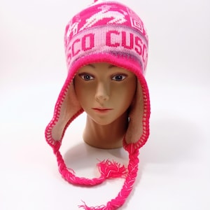 Könnte beinhalten: Eine leuchtend pinke Strickmütze mit Ohrenklappen, verziert mit einer weißen Lama-Grafik und dem in Weiß aufgedruckten Schriftzug CUSCO. Die Mütze hat einen Bommel oben, geflochtene Seitenzöpfe aus pinkem und weißem Garn sowie ein hellbeiges Innenfutter.