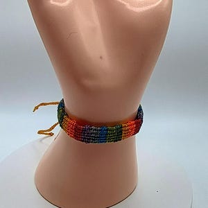 May include: A rainbow-colored friendship bracelet, featuring a woven design with red, orange, yellow, green, blue, and purple threads. The bracelet is tied with two yellow strings. The bracelet is displayed on a mannequin hand.