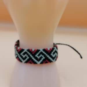 May include: A woven bracelet with a geometric pattern in black, teal, and maroon. The bracelet is wrapped around a light pink form. The bracelet has a black string closure.