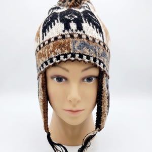 May include: A knit earflap hat featuring a repeating pattern of black, white, and brown geometric shapes and stylized bird motifs. The hat includes long braided tassels hanging from the earflaps, with a textured knit construction throughout.