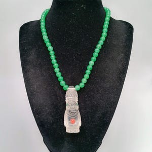 May include: A necklace featuring a strand of round, translucent green beads and a clear, carved quartz pendant shaped like a stylized figure. The pendant includes small green accents at the eyes and a circular red inlay on the chest area.