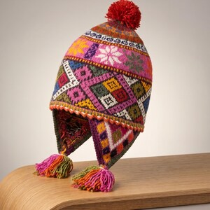 Puede incluir: Un gorro de punto con orejeras que presenta un vibrante patrón geométrico multicolor en tonos rosa, verde, amarillo, azul y blanco. El gorro está rematado con un pompón de lana roja e incluye dos orejeras trenzadas terminadas con borlas.