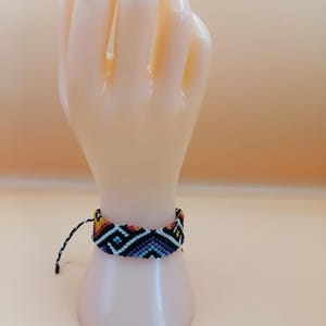 Könnte beinhalten: Ein gewebtes Freundschaftsarmband mit geometrischem Muster. Es zeigt schwarze, blaue, gelbe und rote Fäden. Das Armband wird auf einem Mannequin-Hand präsentiert. Ca. 2 cm breit.