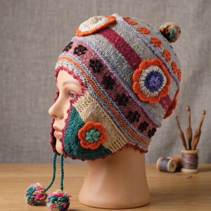 Può includere: Un cappello all'uncinetto multicolore con paraorecchie, base grigia e strisce orizzontali rosa e rosso scuro. Il cappello è decorato con applicazioni floreali arancioni, viola e verdi e include due lunghi lacci intrecciati con pon pon.