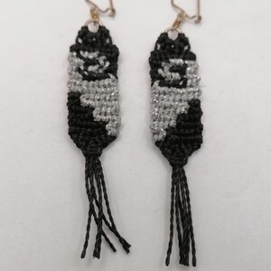 May include: A pair of dangling earrings featuring a rectangular macrame pattern in black and metallic silver thread. Each earring is suspended from a silver-toned metal hook and finishes with a black cord tassel at the bottom edge of the design.