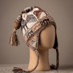 May include: A knit earflap hat featuring a brown, cream, and grey geometric and animal pattern. The hat includes a pointed top with a decorative tassel, two braided side ties with matching tassels, and a thick, textured knit construction.