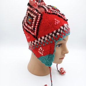 May include: A red knitted earflap hat featuring a multi-coloured geometric pattern in shades of teal, white, and dark brown. The hat includes a pom-pom on the top and two braided tassels with pom-poms hanging from the earflaps, all made of yarn.