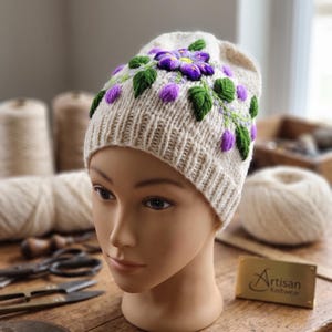 Puede incluir: Un gorro de punto color crema con flores moradas y hojas verdes bordadas. El gorro tiene un borde acanalado y un patrón de punto texturizado. Una pequeña placa rectangular dorada muestra el texto Artisan Knitwear.