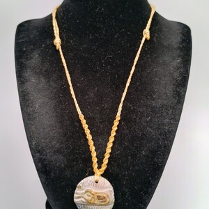 May include: A necklace featuring a round, textured pendant with a metallic finish and a central embossed design, suspended from a light yellow, braided cord with adjustable knots on each side, creating a simple and functional piece of jewelry.