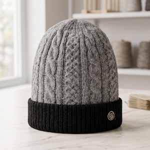 May include: A grey beanie featuring a textured cable knit pattern on the crown and a solid black ribbed cuff. A small, circular gold-toned metal emblem with a stylised logo is attached to the right side of the black cuff.