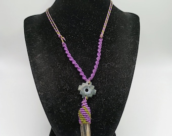 May include: A handmade necklace featuring a clear quartz crystal pendant wrapped in green and purple macrame. The necklace has a purple cord and a gray gear-shaped charm. The pendant is displayed on a black mannequin.