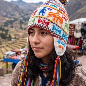May include: A cream-coloured knit chullo hat featuring colourful geometric and animal patterns in red, orange, blue, and green. The hat includes ear flaps, long braided tassels with multi-coloured yarn ends, and a small pom-pom on the top.