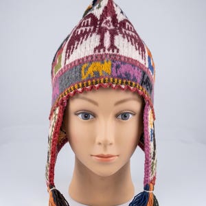 May include: A pointed knit hat featuring a multi-coloured pattern with a prominent bird motif in maroon and white. The hat includes ear flaps with braided tassels at the ends, incorporating shades of blue, orange, and brown throughout the design.
