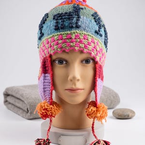 May include: A colorful knit earflap hat featuring a repeating dark blue llama pattern on a light blue background. The hat includes a pink and green checkered band, long braided side ties, and orange and multi-colored yarn pom-poms at the ends.
