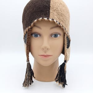May include: A knit earflap hat featuring a split-color design with one side in dark brown and the other in tan. The hat includes earflaps with braided tassels at the ends, incorporating black, brown, and tan yarn, and a white stitched trim.