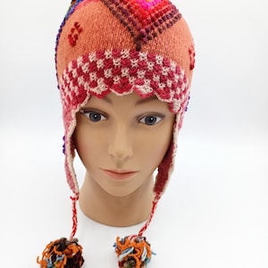 May include: A multi-coloured knit earflap hat featuring a peach-toned base with a geometric diamond pattern in shades of red, pink, and orange. The hat includes a scalloped white and red border, side earflaps, and two dangling braided tassels.