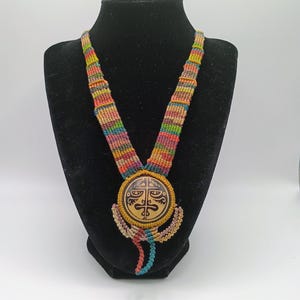 May include: A colorful macrame necklace with a large, round pendant. The pendant features a carved design in shades of brown and tan. The necklace's cord is a mix of rainbow colors, including red, yellow, green, and blue. The necklace is displayed on a black mannequin.