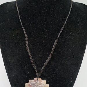 May include: A beige pendant necklace with a black cord. The pendant is a carved, geometric shape with a central hole and detailed relief carvings of birds and other figures. The necklace is displayed on a black mannequin.