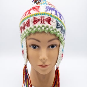 Peut inclure: Un bonnet en tricot blanc avec des oreillettes présentant un motif géométrique coloré en rouge, violet, bleu et vert. Le bonnet comprend un petit pompon sur le dessus et deux pompons tressés avec des fils multicolores aux extrémités.