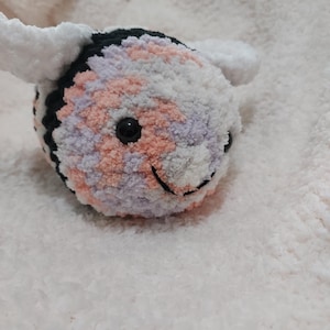 May include: A handmade plush bee with a black head, white wings, and a body in shades of pink, purple, and white. The bee has a smiling face with a black eye and a stitched mouth. The plush toy is made of soft, fuzzy material.
