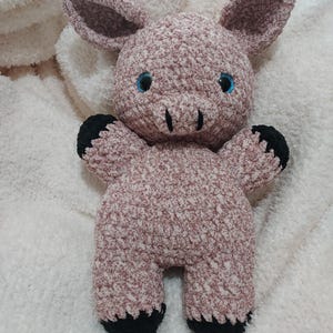 May include: A handmade plush pig toy in shades of pink and white, with black hooves and blue eyes. The pig has large ears and a textured, fuzzy appearance, suggesting a soft, cuddly design. The toy is likely intended for children.