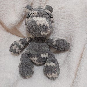 May include: A handmade, gray and white crochet hippo plush toy. The hippo has small ears, closed eyes, and a rounded body with arms and legs. The plush is made of soft, textured yarn and is resting on a white, fluffy surface.
