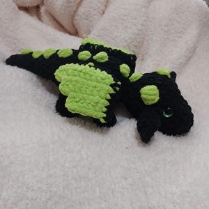 May include: A handmade crochet dragon in black and lime green. The dragon has green wings and spots, with a long tail and small horns. The dragon has green eyes and is lying on a white, fluffy surface.