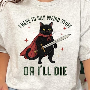 May include: Light grey t-shirt featuring a black cat wearing a red cape and holding a sword. The text on the shirt reads "I HAVE TO SAY WEIRD STUFF OR I'LL DIE". The design includes red star accents.