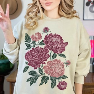 Puede incluir: Sudadera color crema con un diseño floral de peonías burdeos y rosas y hojas verdes. El gráfico está centrado en la parte delantera de la parte superior de manga larga. El estilo general es informal y relajado.