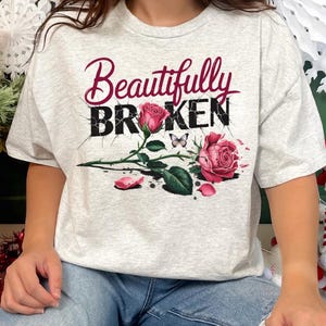 May include: Light gray t-shirt with the words "Beautifully Broken" in burgundy script and black block letters. The design includes two pink roses, a butterfly, and rose petals.
