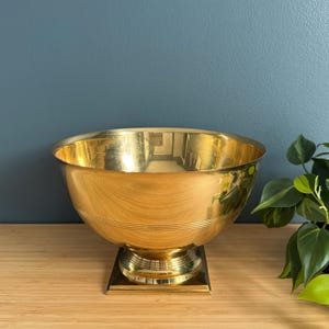 May include: A large, gold-colored metal bowl with a wide, rounded shape. The bowl sits on a square base and has a decorative, ridged band around the middle. The interior reflects light, and the overall design is vintage.