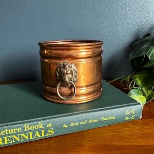 May include: A cylindrical copper planter featuring a brass lion head ornament with a ring handle. The planter rests on a dark green book titled Picture Book of Perennials by Arno and Irene Nehrling, with visible gold lettering on the spine.