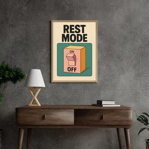 Puede incluir: Un estampado enmarcado con las palabras "REST MODE" sobre un gráfico de un interruptor con las etiquetas "ON" y "OFF". El interruptor es naranja sobre un fondo verde azulado. El estampado se muestra en una consola de madera oscura con una lámpara y vegetación.