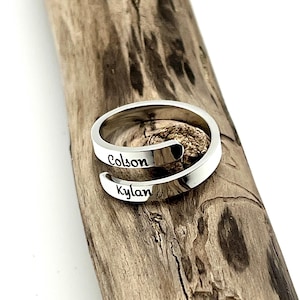 Puede incluir: Un anillo envolvente plateado con los nombres "Colson" y "Kylan" grabados. El anillo se muestra sobre un trozo de madera flotante, mostrando su diseño y los nombres grabados.