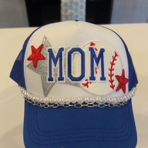 May include: A blue and white trucker hat featuring the word MOM in blue letters. The design includes a silver glitter star, a baseball graphic with red stitching, two red embroidered stars, a string of white pearls, and a silver chain.