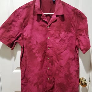 May include: A short-sleeved, button-down shirt in a deep burgundy color, hanging on a white hanger. The shirt has a single chest pocket and wooden buttons. The fabric has a subtle, textured pattern.