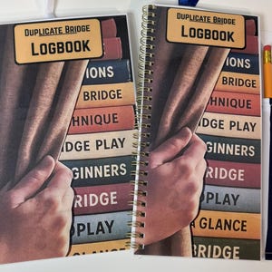 Può includere: Due quaderni Duplicate Bridge rilegati a spirale. Ognuno presenta un design di copertina con una pila di libri colorati e il titolo "Duplicate Bridge Logbook". Uno ha una matita in una tasca a righe.