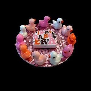 以下が含まれることがあります： ピンクのグリッタートレイに、ピンク、オレンジ、ブラウンの「In My Dance Era」のサインが中央に置かれています。トレイは、ピンク、オレンジ、ホワイトの小さなゴム製のアヒル人形10個の円形に囲まれています。