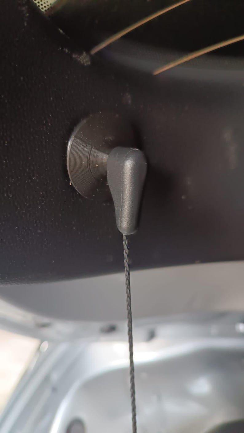 May include: A close-up of a black car interior component. A black plastic pull handle with a string is attached to a circular base. The handle is teardrop-shaped and hangs down.