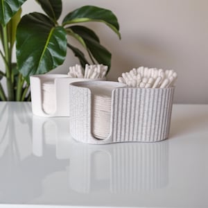 Può includere: Due contenitori in ceramica, uno bianco e uno grigio maculato, contengono dischetti di cotone e cotton fioc. I contenitori hanno un'apertura curva per un facile accesso. Una pianta verde è visibile sullo sfondo.