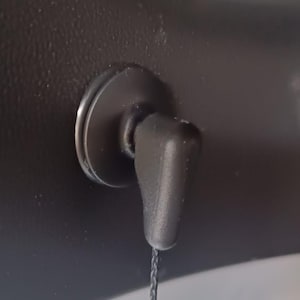 May include: A black plastic car interior component with a circular base and a small, angled handle. A thin, dark cord hangs from the handle. The component is mounted on a dark, textured surface.