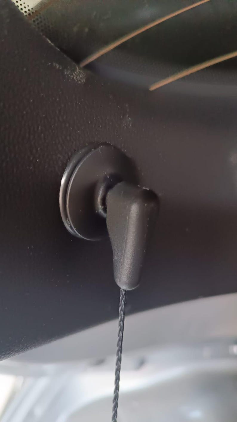 May include: A black plastic car interior component with a circular base and a small, angled handle. A thin, dark cord hangs from the handle. The component is mounted on a dark, textured surface.
