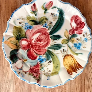 Puede incluir: Un plato de cerámica blanco con borde festoneado y ribete azul. El plato está decorado con flores pintadas a mano en tonos rojos, rosas, amarillos y azules, con hojas verdes. El diseño floral está centrado en el plato.