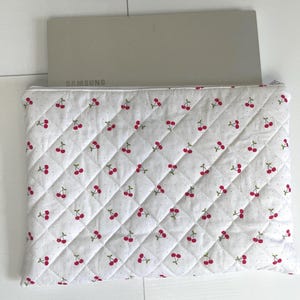 Puede incluir: Funda acolchada blanca para portátil con un estampado de cerezas rojas y tallos verdes. Dentro hay un portátil gris. El nombre de la marca "SAMSUNG" está impreso en el portátil.